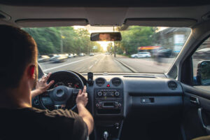Imagem de dentro de um carro mostrando, de costas pessoa dirigindo. Destacando o painel do carro e visão da rua à frente.